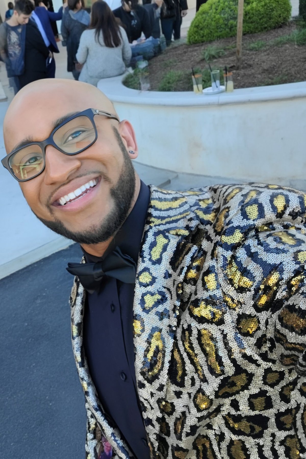 Man wearing glasses and a sequined animal print jacket smiles at the camera at an outdoor event