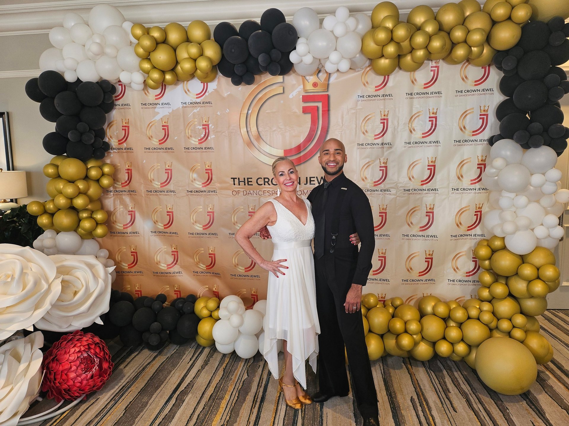 Two people posing at an event backdrop decorated with gold, black, and white balloons and large floral arrangements