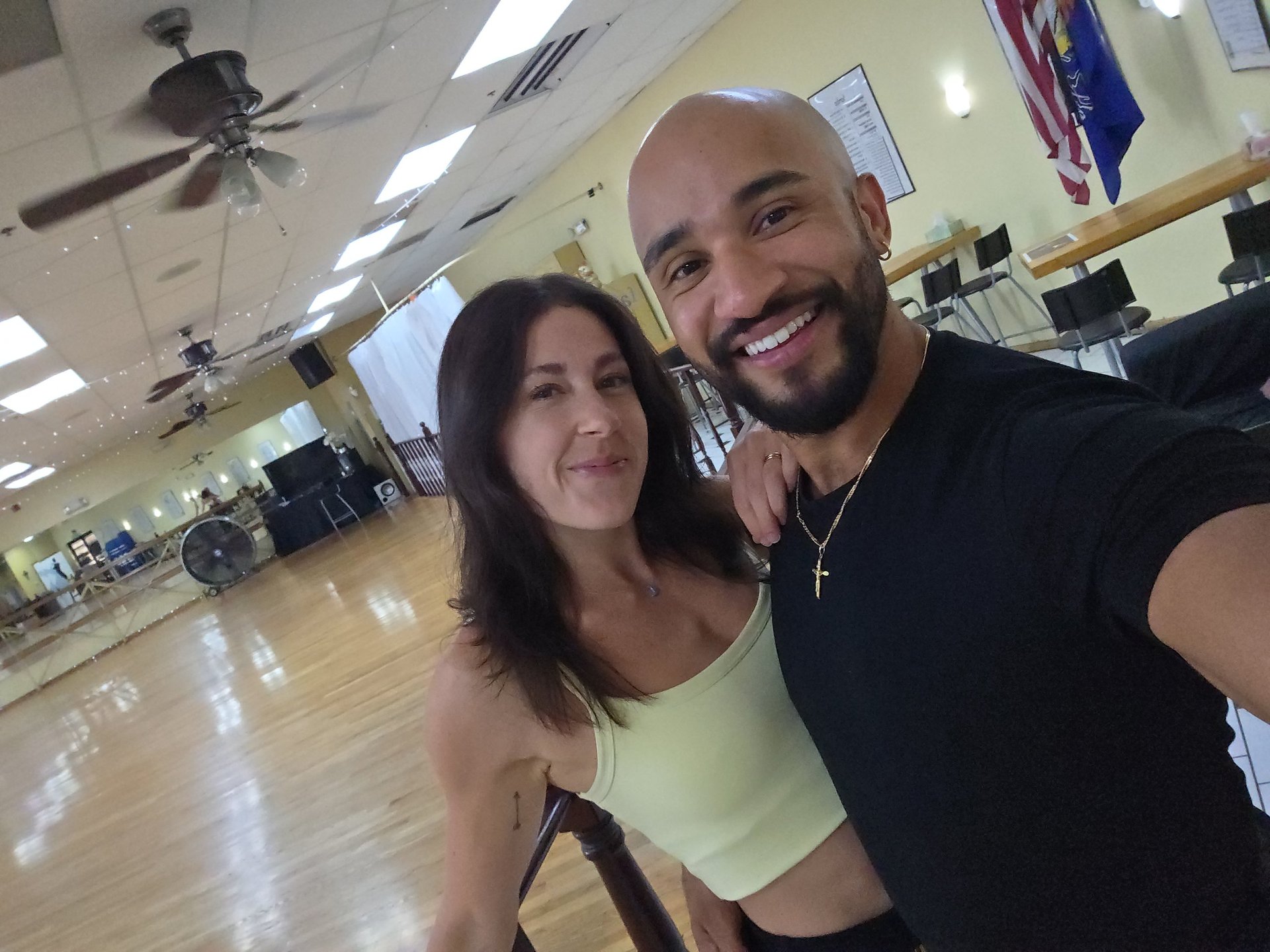 Two people smiling at the camera inside a fitness studio with wooden floors and ceiling fans