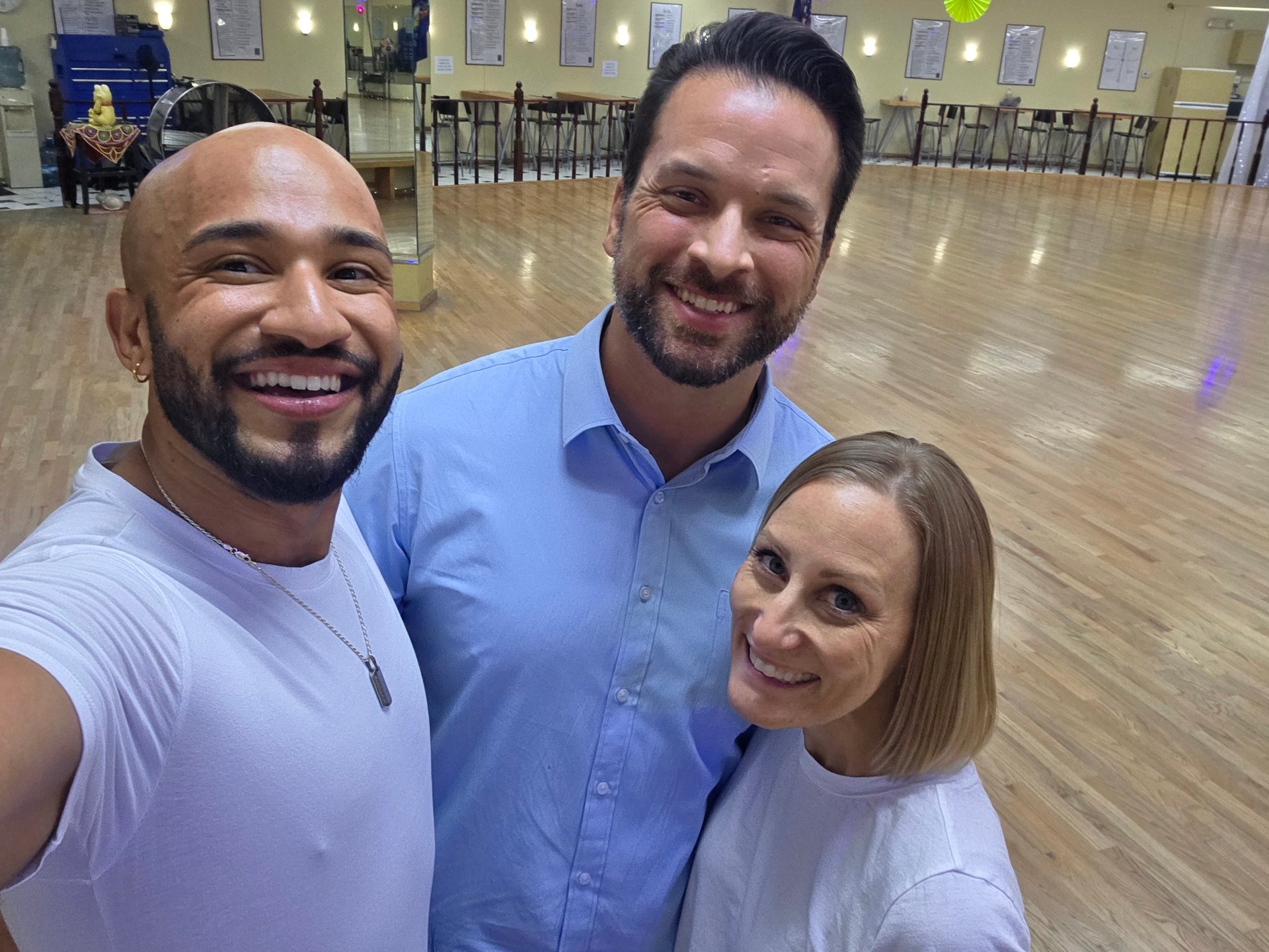 Three smiling people posing together in an indoor gym or community center with wooden flooring