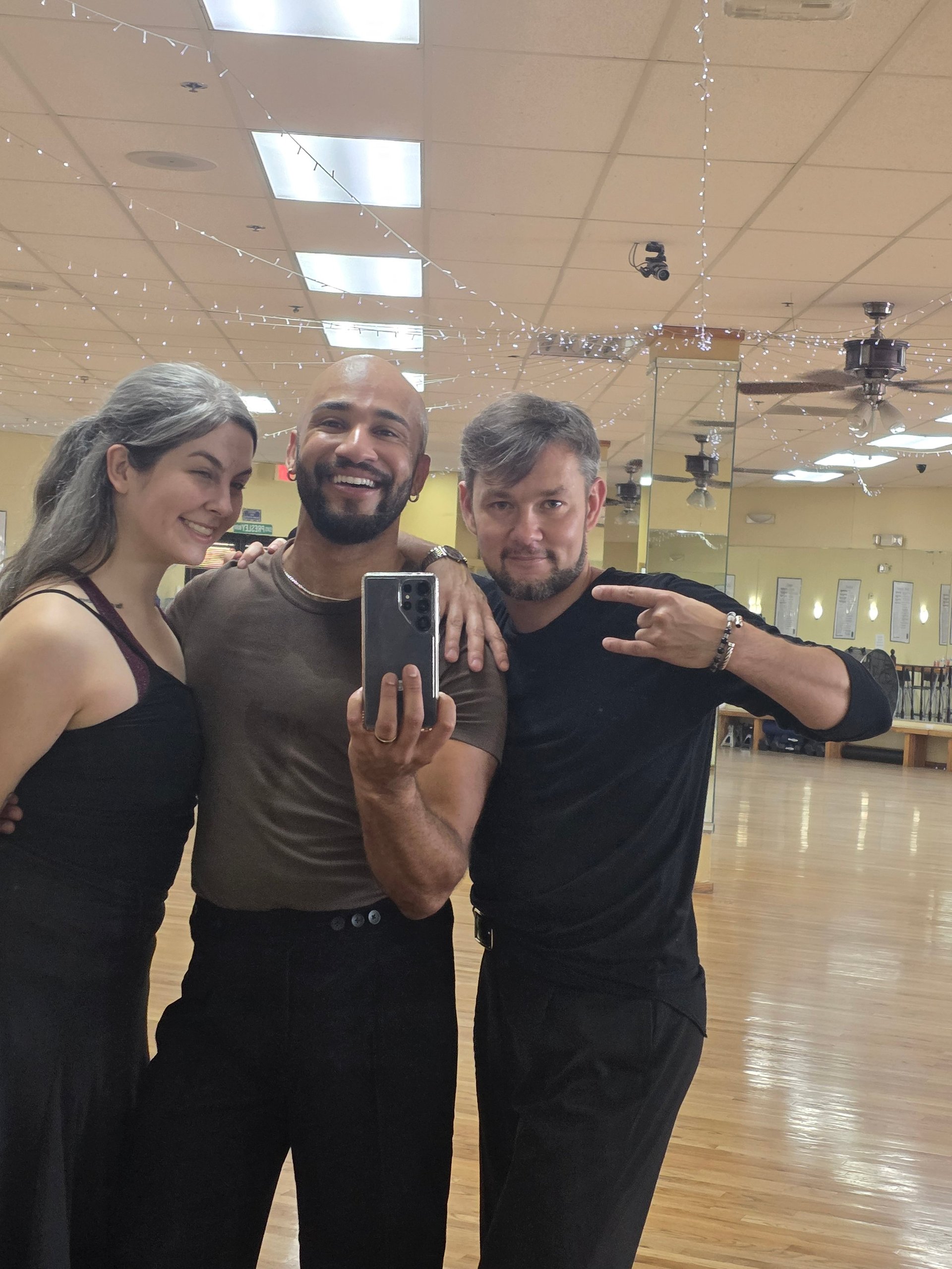 Three people taking a selfie in a dance studio with wooden floors and bright ceiling lights