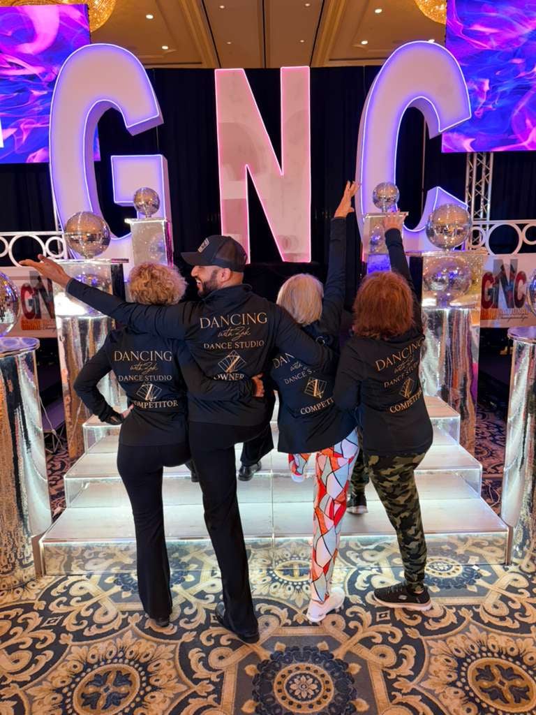 Four people posing together at a GNC event with large blue letters and purple lighting in a decorated ballroom