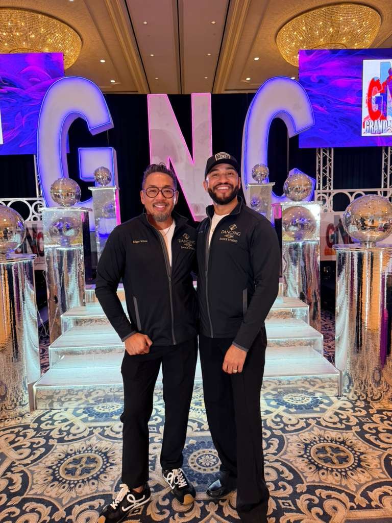 Four people in black jackets stand facing a large illuminated GNO sign at an indoor event venue with colorful stage lighting
