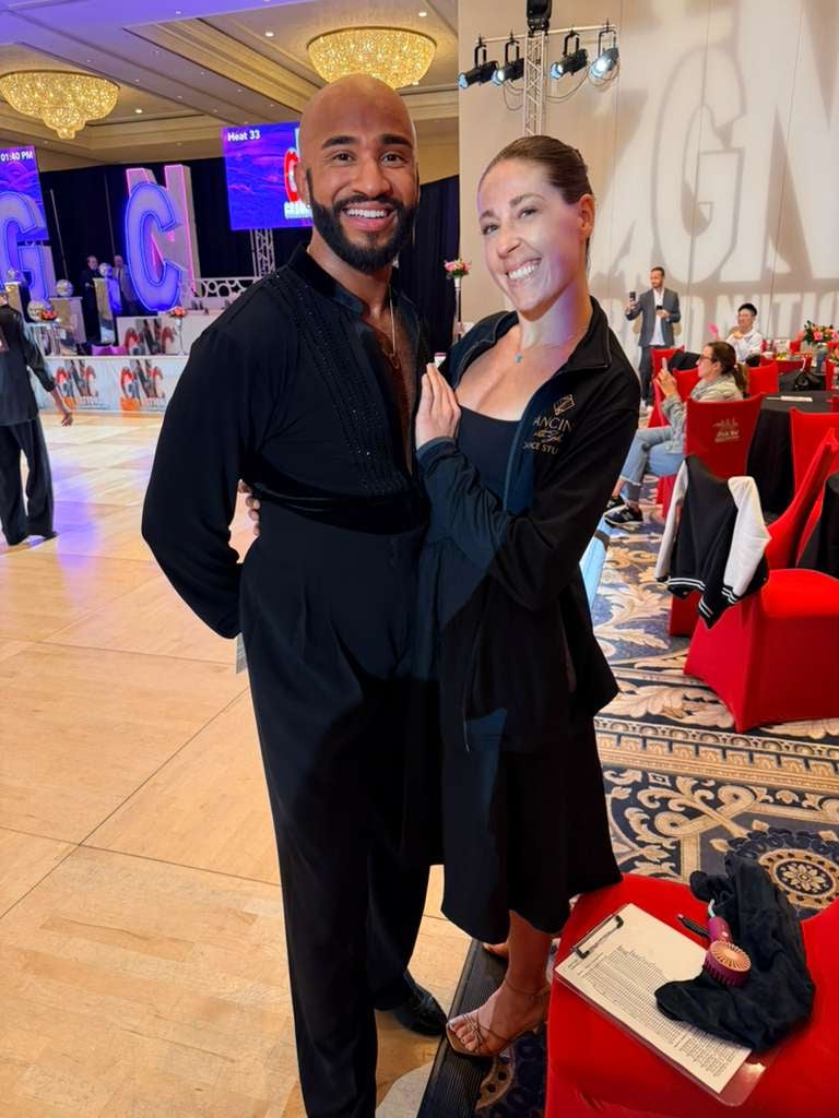 Two people in black attire posing together at an elegant ballroom event with red-clothed tables and ornate ceiling lighting