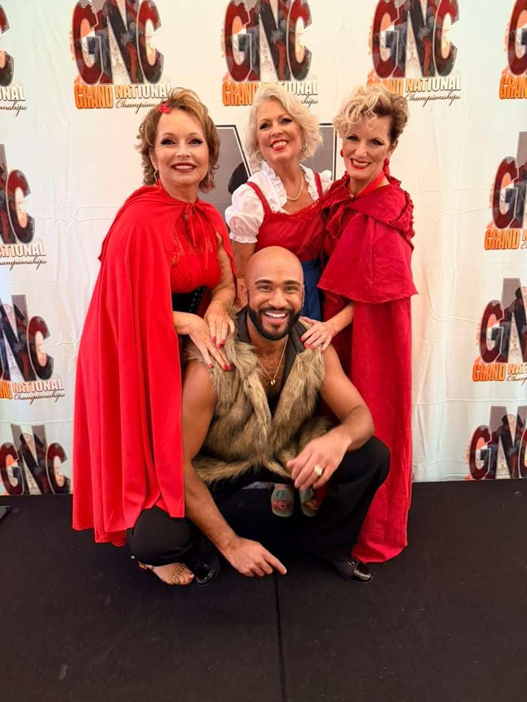 Four people posing together at a GNC branded event, with three women in red capes and one man in a tan vest crouching in front